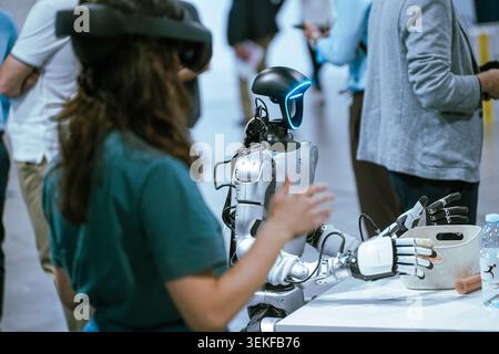 A young woman wearing a virtual reality headset controls a humanoid robot during a demonstration at an event. Stock Photo