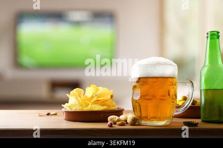 Cold beer mug with chips and snacks on a wooden table while a football match plays blurred in the background at home. Stock Photo