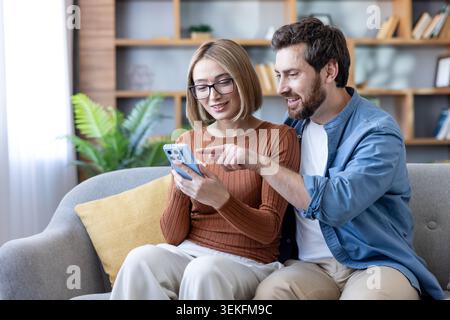 Young couple sitting on a sofa at home, man pointing to a mobile phone screen held by woman, sharing online content, browsing app, and enjoying digita Stock Photo