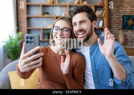 Young smiling couple at home communicating with friends or family via video call, actively waving hands during online conversation, showing connection Stock Photo