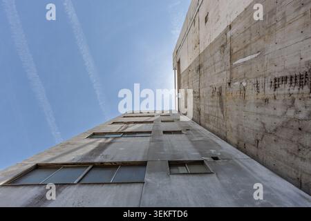 Stark, low-angle view up raw, brutalist concrete walls jutting into a clear blue sky, marked with faint jet trails and hinting at urban solitude. Stock Photo