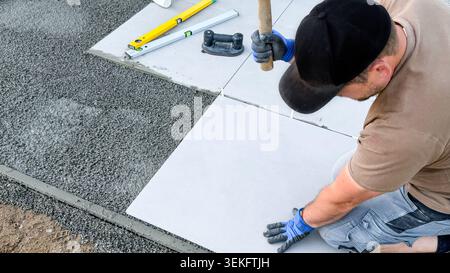 Male construction worker laying concrete tiles on gravel surface using a hammer and level tool, with additional tools visible in the background Stock Photo