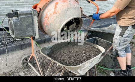 Construction worker using concrete mixer to pour gravel into wheelbarrow on building site, with gray brick wall and tools visible in the background Stock Photo