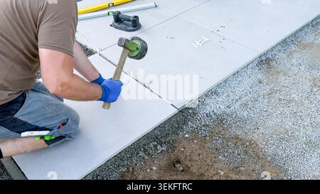 Male worker laying large concrete tiles on gravel surface using a rubber mallet, tools and measuring equipment visible in the construction area Stock Photo