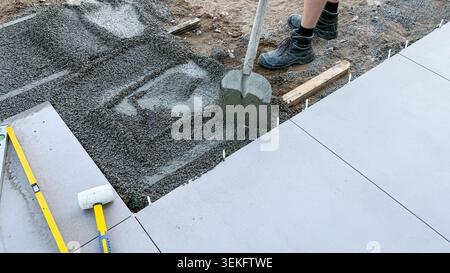 Construction worker using a shovel to spread wet concrete on a newly laid floor surface with tools and wooden supports visible in the outdoor setting Stock Photo