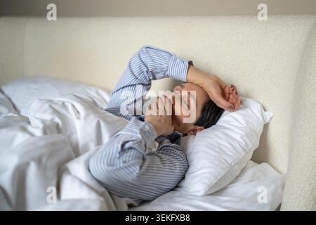Dreamy senior woman comfortably lying in bed covered in cozy blanket and yawning after waking up in morning, sleepy lazy elderly female awakening at early hours after good night rest and sweet dream. Stock Photo