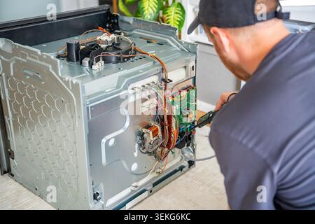 Technician testing electronic components and wiring inside disassembled electric oven. Appliance diagnostics and professional repair service concept Stock Photo