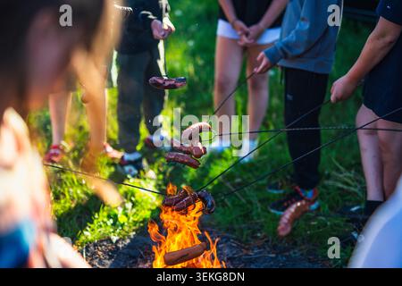 A close-up shot of several sausages being grilled over open flames by a group of friends outdoors Stock Photo