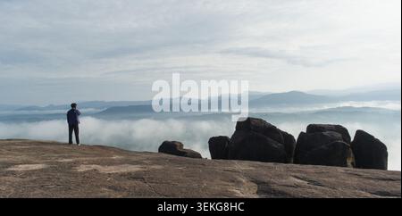 A wide landscape shot featuring a single person standing on a large, flat rock formation, gazing out over a vast valley filled with thick morning mist Stock Photo