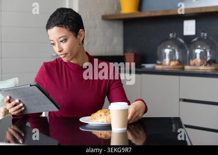African American woman sitting at black table in cafe in burgundy top using tablet with croissant Stock Photo
