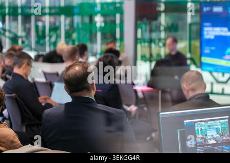 Business audience attending conference presentation in modern glass meeting room, laptops and screens visible, focused professional atmosphere. Stock Photo