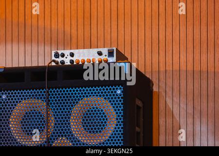 Warm-lit close-up of a vintage amplifier head sitting on a perforated speaker cabinet against wooden wall, showing control knobs, cable connection and Stock Photo