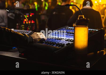 Experienced audio technician operates a mixing console during a live concert, adjusting faders and levels under stage lighting while a glowing water b Stock Photo