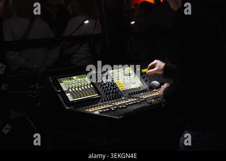 Live sound engineer adjusts a digital mixing console at a crowded concert, balancing levels on illuminated touchscreens and faders while holding a mic Stock Photo