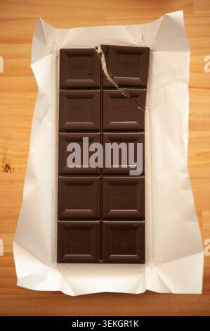 Open dark chocolate bar in white wrapper on wooden table, one broken piece, minimalist food still life Stock Photo