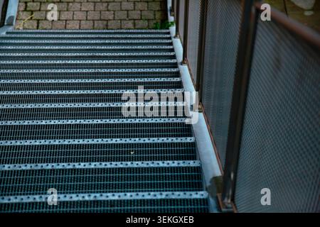 This professional outdoor photograph captures a modern industrial metal staircase looking from a high angle perspective. The camera focuses on the long steel grating steps designed with a non slip Stock Photo