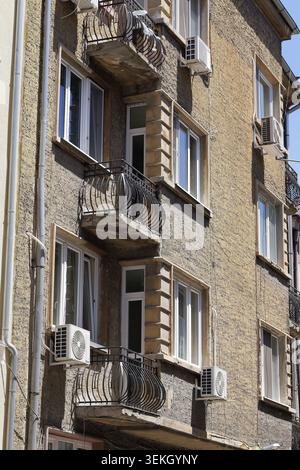 Old residential building facade displaying multiple balconies with ornate iron railings, windows, and outdoor air conditioning units on a sunny day in Stock Photo