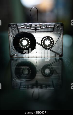 Transparent compact cassette tape standing upright with magnetic tape pulled out on reflective dark surface in moody atmospheric dark background Stock Photo