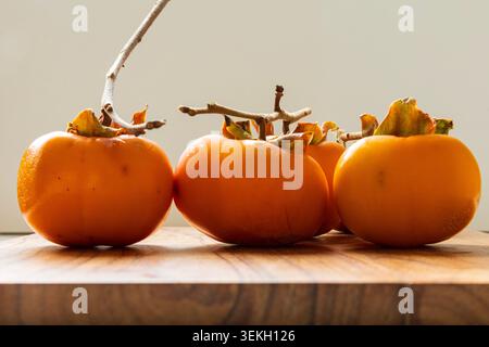 Three ripe persimmons arranged on a wooden cutting board in soft natural light. Seasonal autumn fruit with warm colors and minimalist composition. Stock Photo