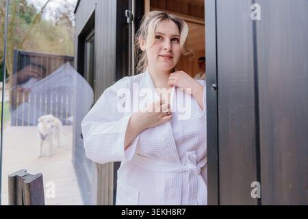 Calm Young White Woman Adjusting Bathrobe At Sauna Door Stock Photo
