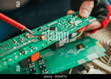 Close-up of a technician using a soldering iron to repair a burnt and damaged electronic circuit board, showcasing the intricate components and the precision required in electronics repair Stock Photo