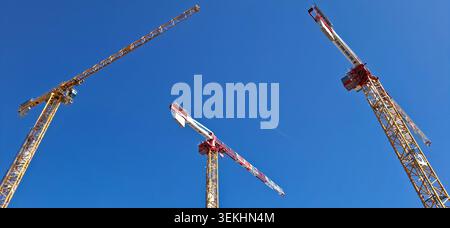 Low angle view of three industrial construction cranes against a clear blue sky with an airplane contrail in the center, France. Stock Photo