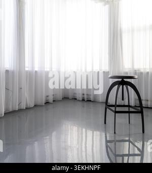 view of a small black stool on a shiny floor in front of windows with white curtains Stock Photo