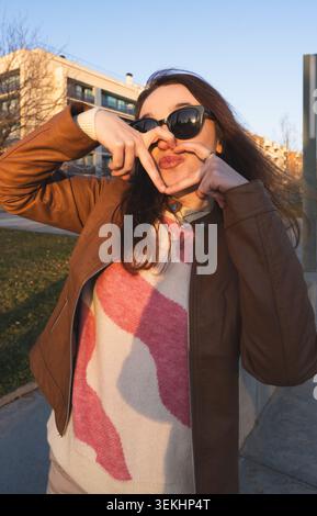 Young woman smiling and making a heart shape with her hands outdoors at sunset Stock Photo