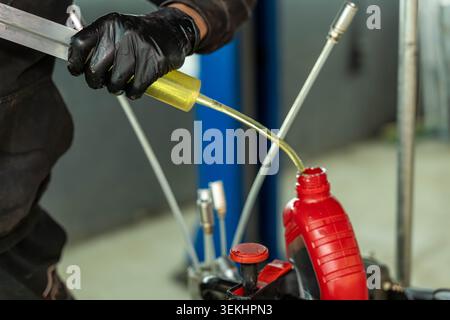 A close up view of a worker wearing black gloves pouring yellow liquid into a red plastic container in an industrial workshop, with blurred tools Stock Photo