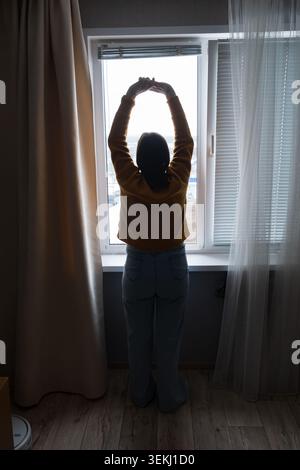Pregnant woman standing by a window in soft morning light, hands on belly indoors Stock Photo