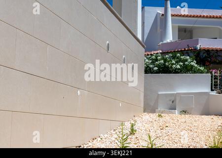 A textured beige stone facade with horizontal lines and circular vents is contrasted by whitewashed buildings and lush greenery under a clear blue sky Stock Photo