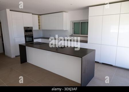 A spacious, contemporary kitchen featuring high-gloss white cabinetry, a dark stone island with a built-in hob, and stainless steel appliances Stock Photo