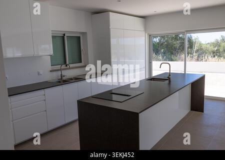 A contemporary kitchen featuring high-gloss white cabinetry, a dark countertop island with an integrated cooktop, and a large sliding glass door leadi Stock Photo