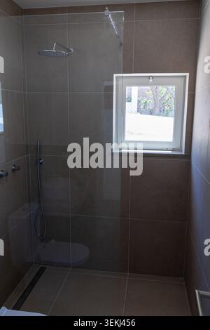 A contemporary bathroom features a spacious walk-in shower with a rainfall showerhead and a handheld sprayer, complemented by a small window offering Stock Photo