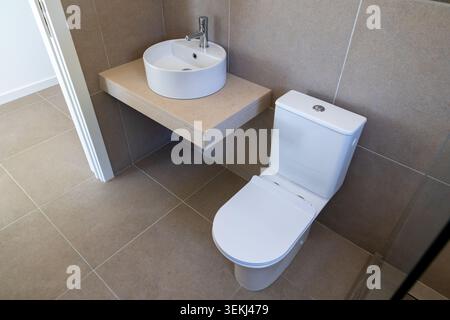 A clean and contemporary bathroom featuring a white ceramic toilet and a round vessel sink mounted on a beige floating countertop with tiled walls and Stock Photo