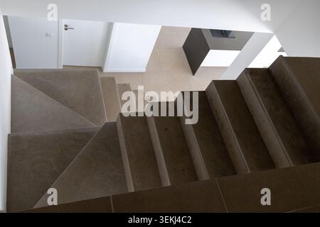 A high-angle view of a contemporary concrete staircase with sharp angles and clean lines, leading to a bright hallway with doors Stock Photo