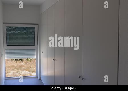 A clean, contemporary room features a floor-to-ceiling white wardrobe with subtle handles and a large window revealing a sun-drenched, arid outdoor sc Stock Photo