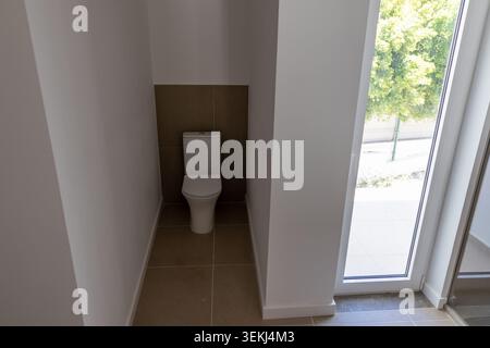 A white ceramic toilet is positioned against a tiled wall in a bright, contemporary bathroom space, offering a view of greenery outside Stock Photo