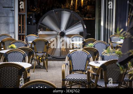 A large industrial fan stands in an empty outdoor area of a cafe between numerous black and white woven chairs and round tables Stock Photo