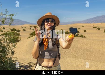 Woman wearing a hat and sunglasses, vlogging her desert adventure for social media, holding an action camera on a selfie stick against the vast death Stock Photo