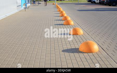 Yellow concrete hemisphere bollards stand on a paved sidewalk. Safety barriers separate the shopping mall parking lot zone. People walk along a paved Stock Photo