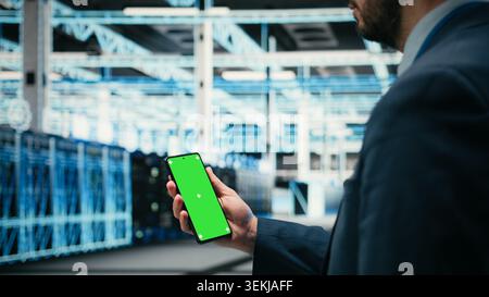 Green screen smartphone used by data center admin managing wireframe mesh gear helping power internet services. Worker uses mockup mobile phone in server farm for digital storage systems, camera A Stock Photo