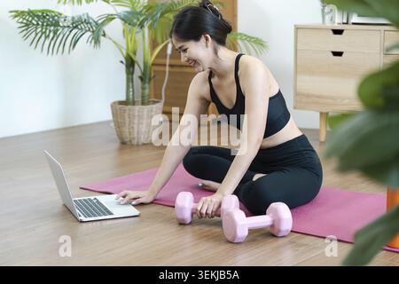 A young woman in activewear practicing yoga at home, using a laptop for guidance, surrounded by plants and fitness equipment Stock Photo