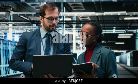 Data center engineering team using AR tech, working together to futureproof company network. Teamworking server room colleagues brainstorm, using devices to increase cybersecurity protection, camera A Stock Photo