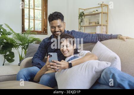 Young couple watching online content in a smart phone sitting on a sofa at home in the living room Stock Photo