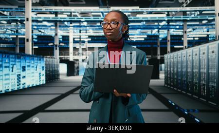 Female technician walking in data center, using laptop and augmented reality to manage database storage rigs. African american woman working in cloud computing server farm, VFX overlay, camera A Stock Photo