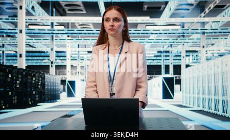 Woman at server hub workplace desk monitors rigs power consumption with laptop. Data center employee oversees equipment with notebook, ensuring industry standards compliance, camera A Stock Photo