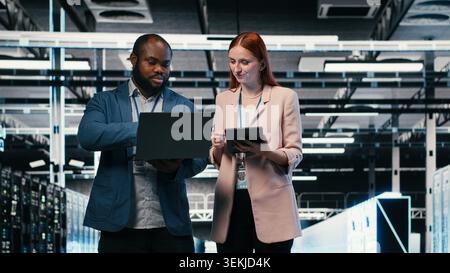Team of server farm software developers using laptop to optimize electronics. Data center teamworking colleagues brainstorming, using notebook to monitor system performance bottlenecks, camera B Stock Photo