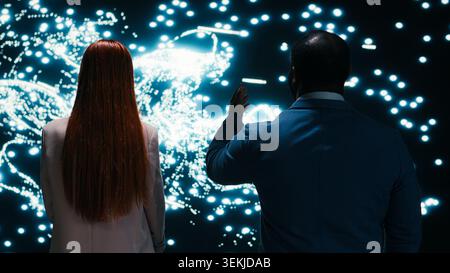 Engineering team monitoring AI model cognitive systems using AR projection, collaborating. RD tech startup coworkers brainstorming, using laptop to study neural network activity, camera B Stock Photo
