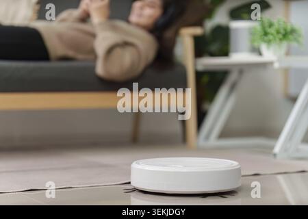 A robot vacuum operating on a tiled floor in a contemporary living room while a person relaxes on a couch in the background Stock Photo